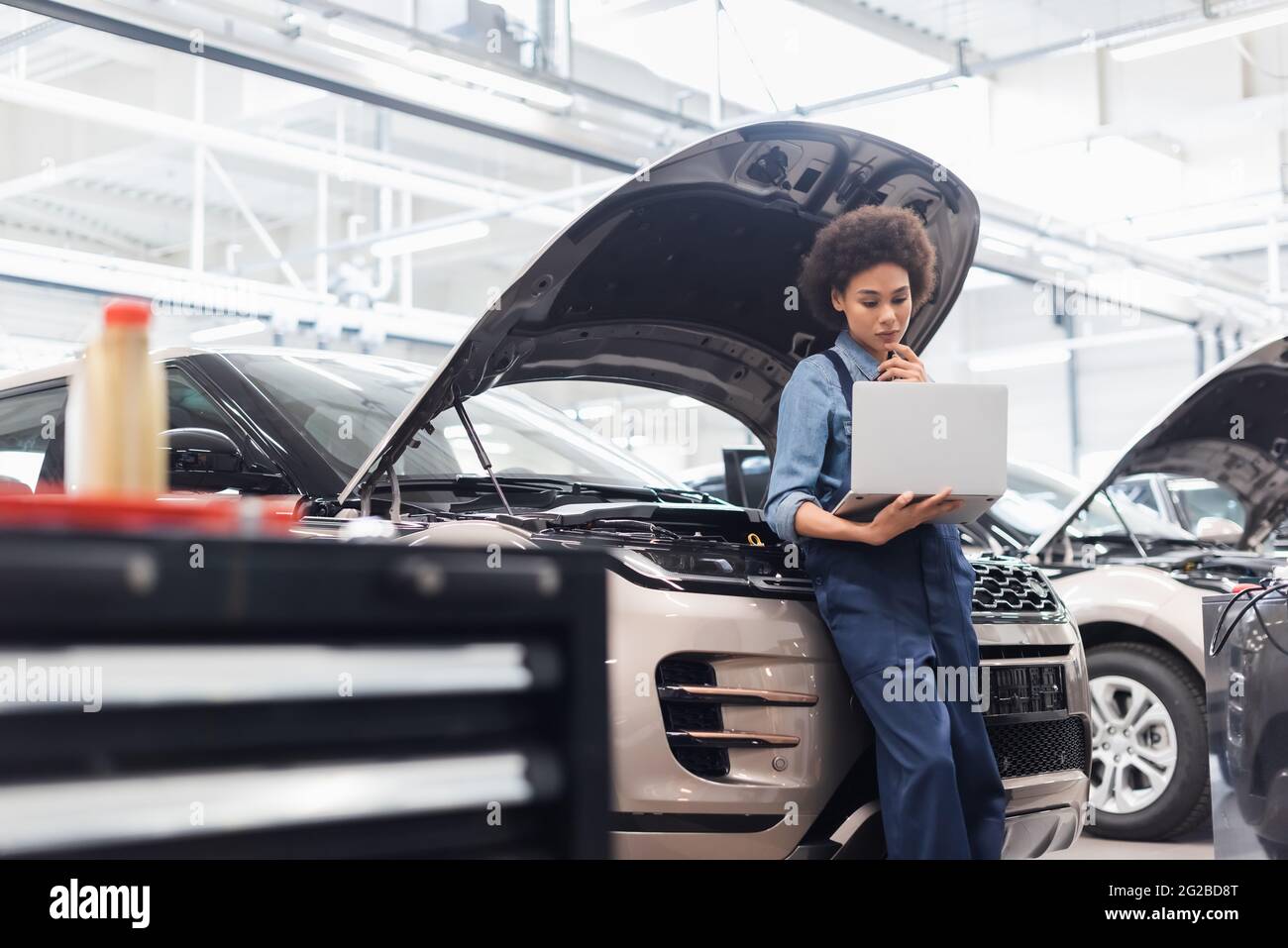 young african american mechanic holding laptop near car with open hood in auto repair service
