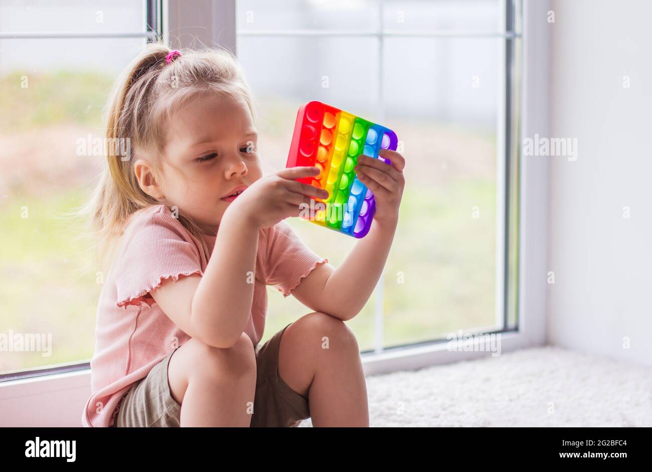 A little blonde girl sits near the window and plays with new trend sensory toy - rainbow pop it. Antistress сolorful toy simple dimple. Squishy soft b Stock Photo