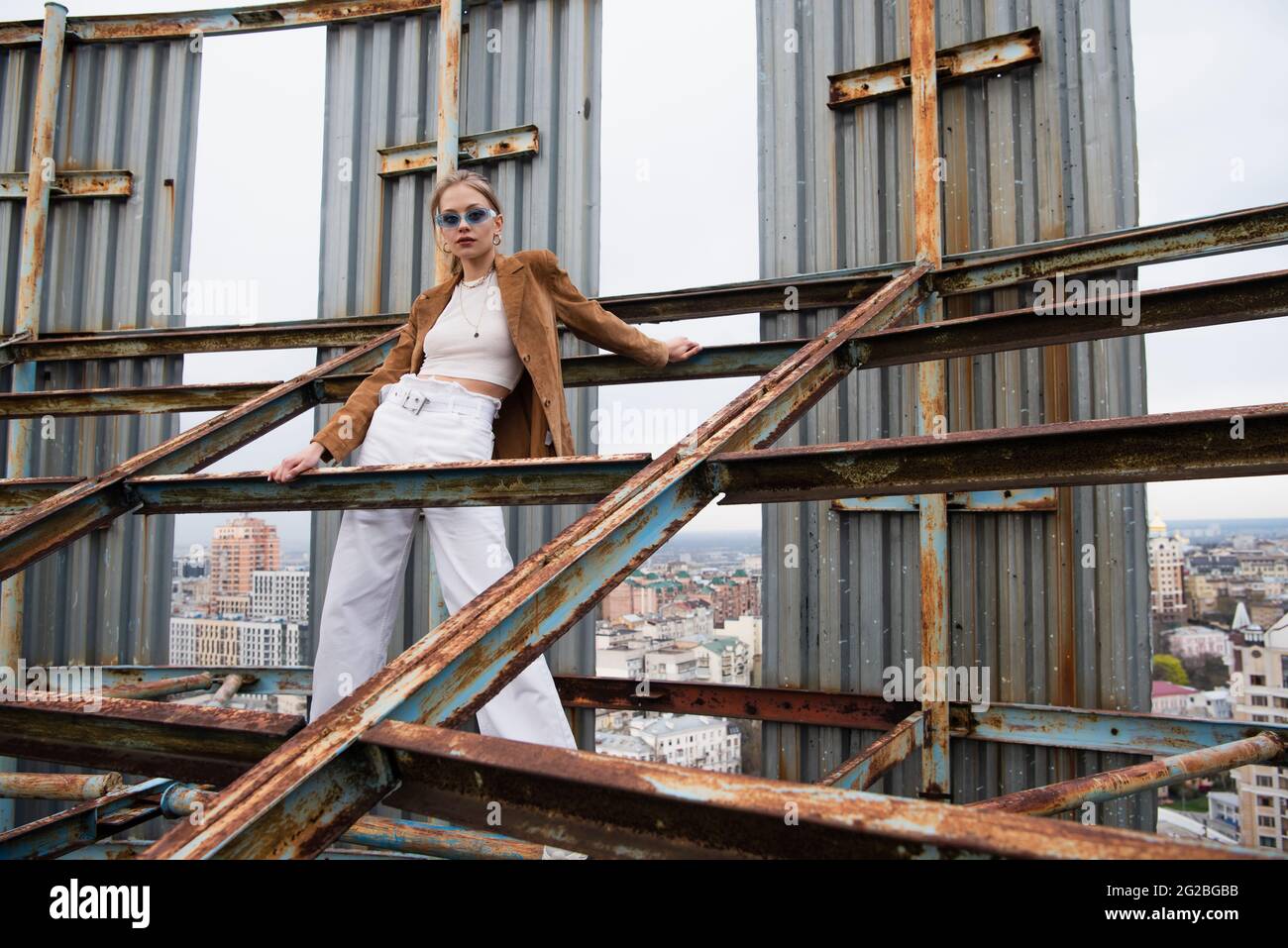 young and pretty model in stylish outfit and sunglasses standing on weathered construction of rooftop Stock Photo