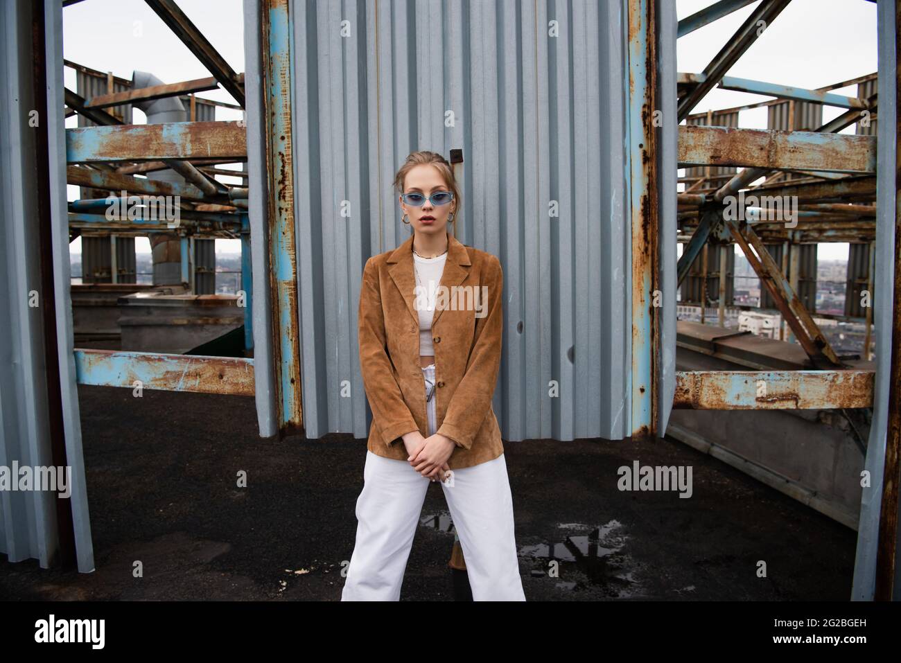 blonde model in blue sunglasses posing near rusty construction on rooftop Stock Photo