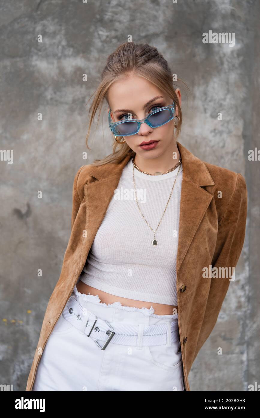 model in blue sunglasses and stylish outfit posing near grey concrete wall Stock Photo