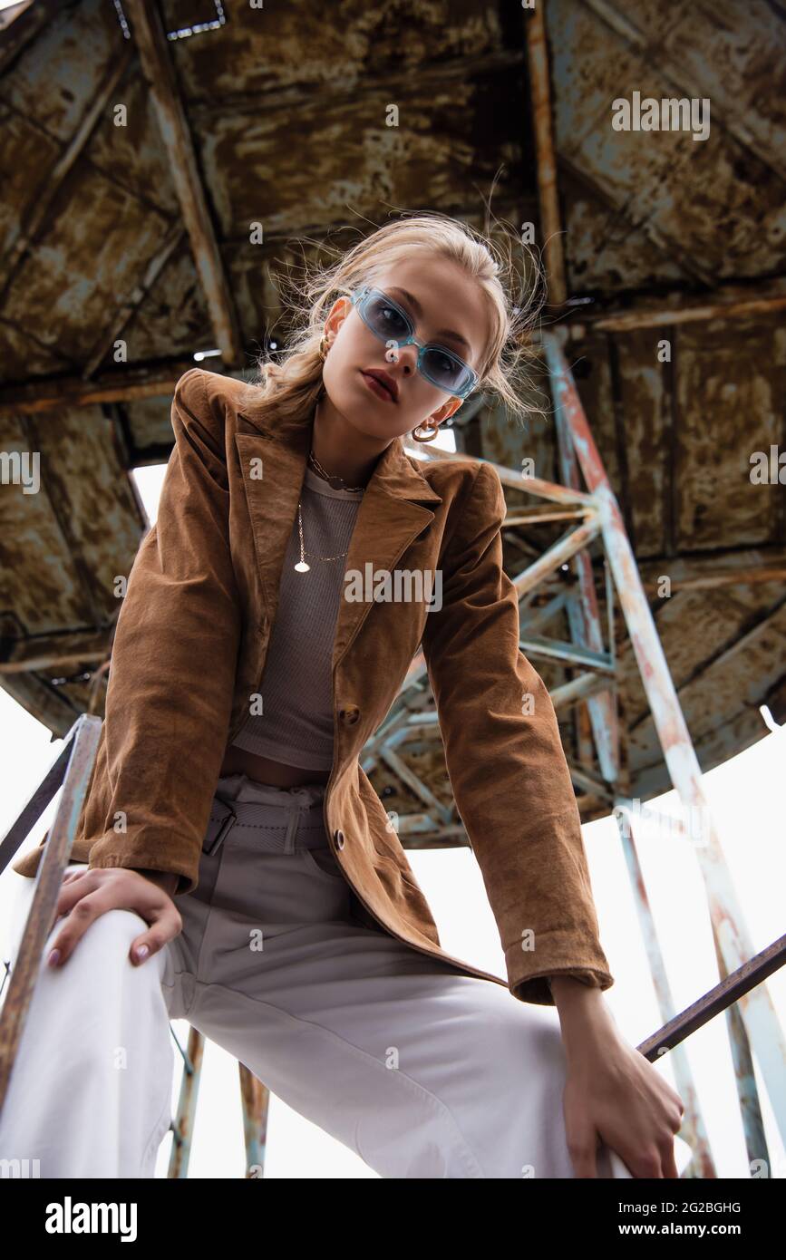 low angle view of pretty model in blue sunglasses and trendy outfit posing near construction on rooftop Stock Photo