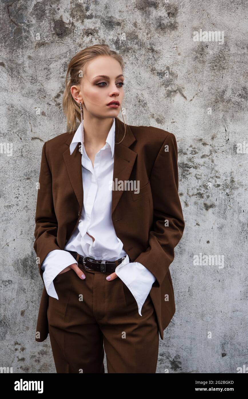 young woman in stylish outfit posing with hands in pockets near ...