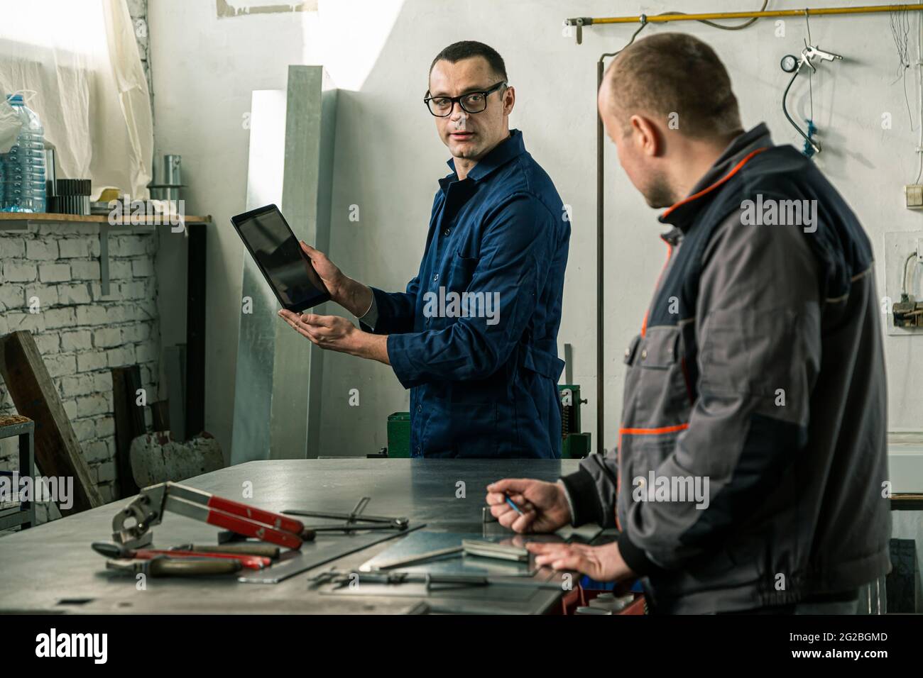 A tinsmith and his fellow colleague engineer are working together on a work task in their workshop, while the engineer is using tablet. Stock Photo