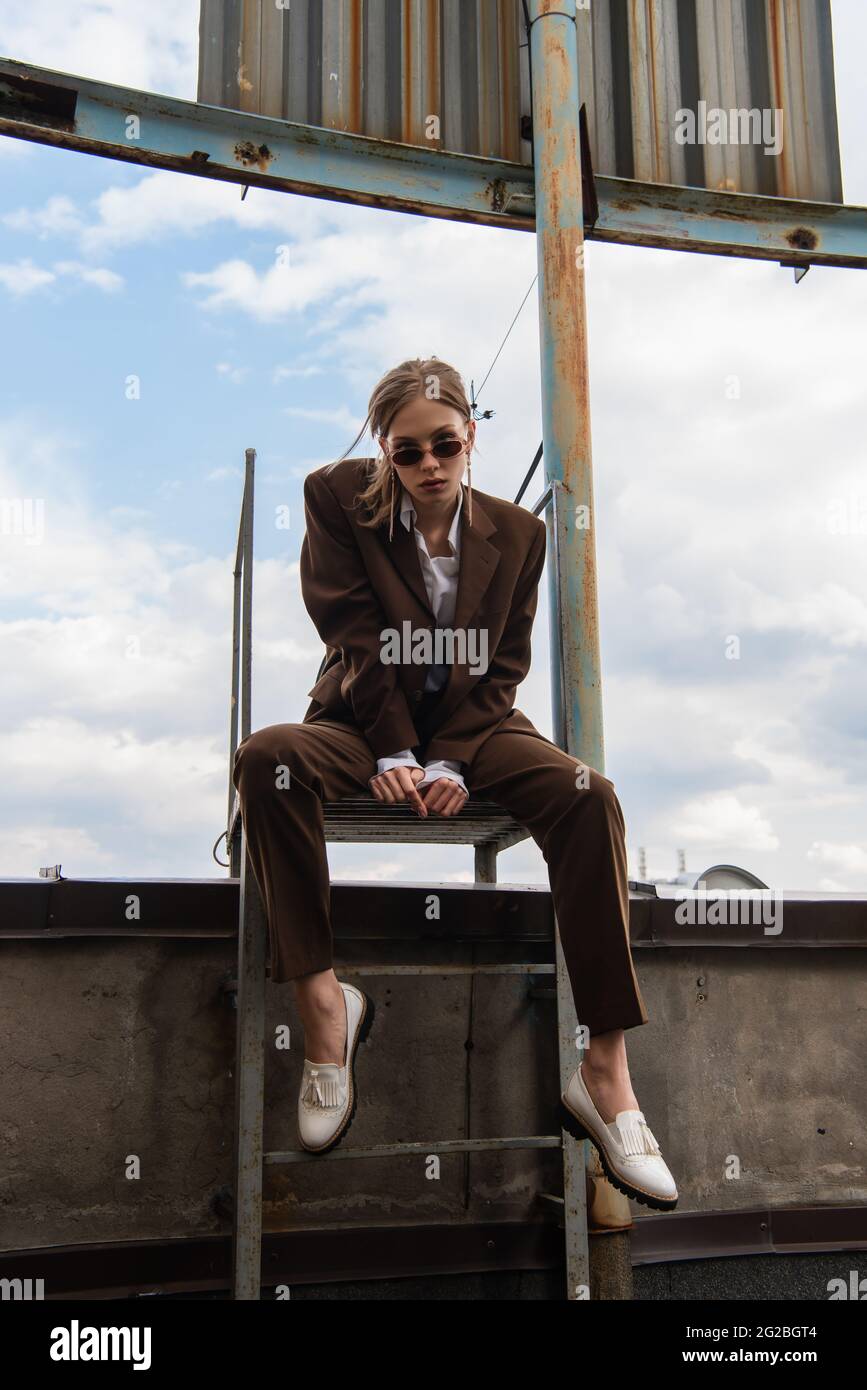 full length of young woman in sunglasses and trendy suit sitting on metallic rusty stairs on rooftop Stock Photo