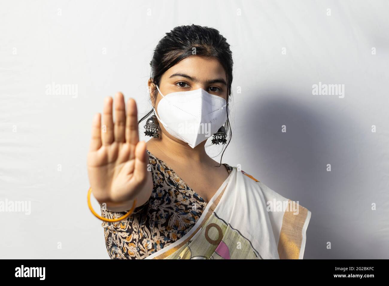 An Indian woman in saree wearing N 95 nose mask showing palm as social distancing on white background Stock Photo