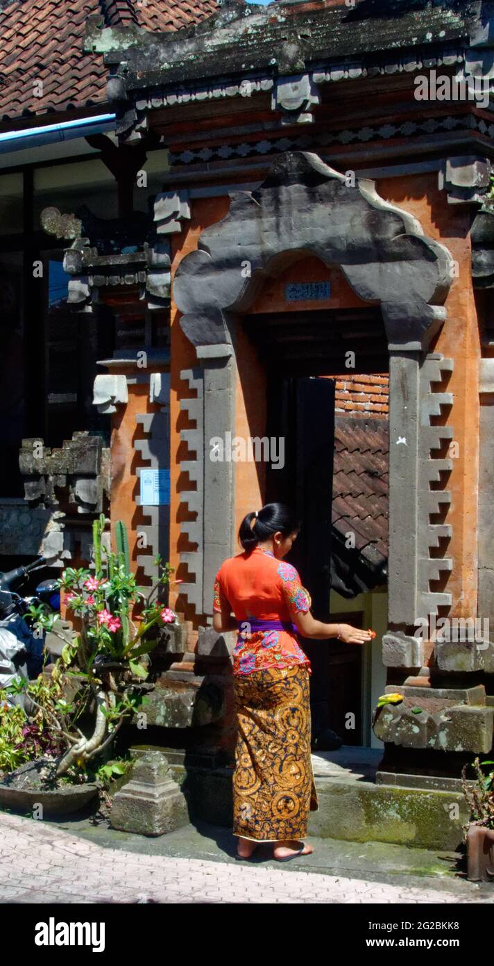 Indonesian woman in traditional dress putting morning offerings outside a house entrance Stock Photo