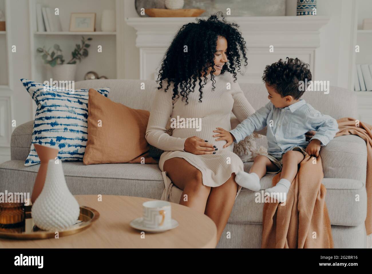 Cute little boy touching belly of his pregnant mom while relaxing together on sofa in living room Stock Photo