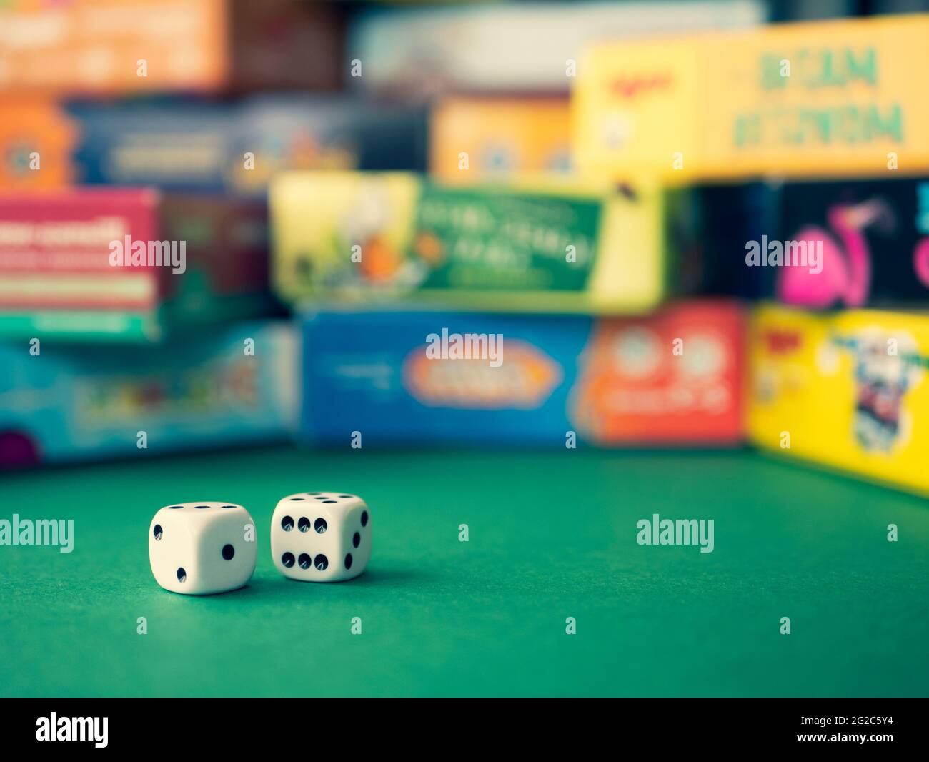 White dice on the green surface on the blurred background of colorful board game boxes. Concept of entertainment under lockdown Stock Photo