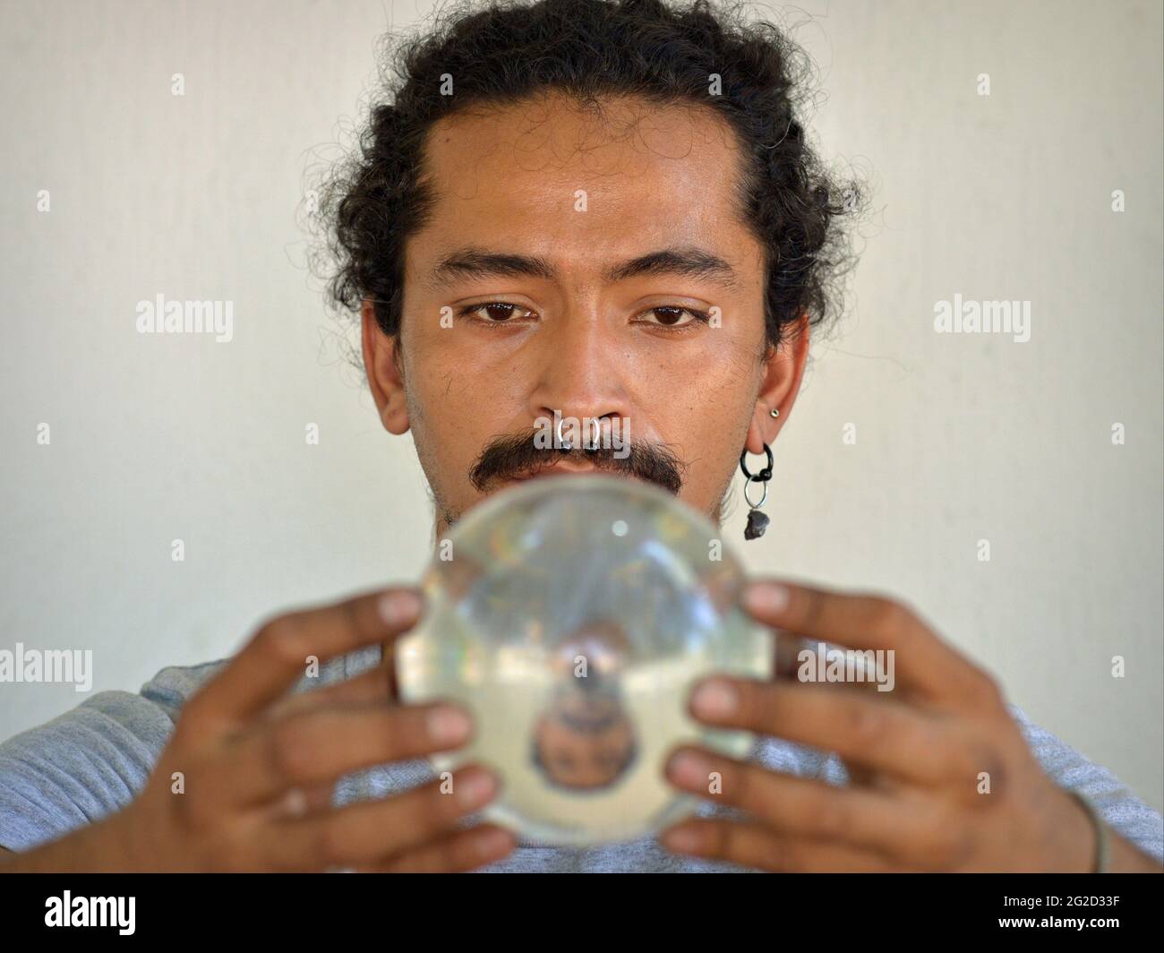 Handsome young Latino man holds a crystal ball with both hands and looks down at the crystal ball and his vertically flipped mirror image. Stock Photo