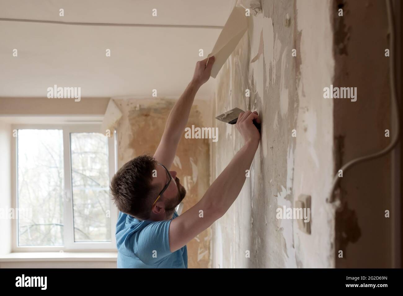 Hardworking man removes old wallpaper from the wall with a spatula in the room. Home repairs. Stock Photo