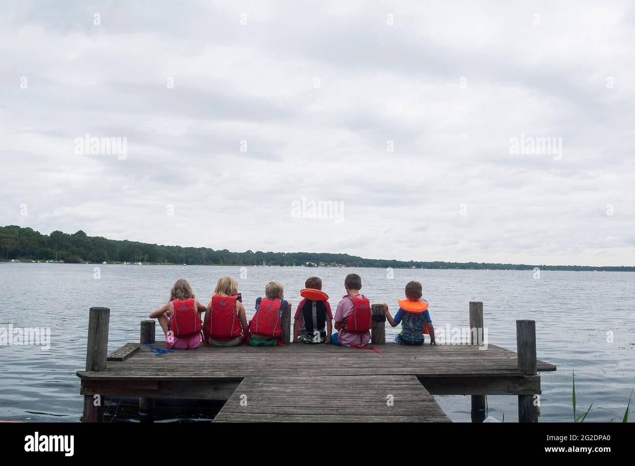 A group of young kids sitting on a wooden dock by a lake, with life jackets on. Stock Photo