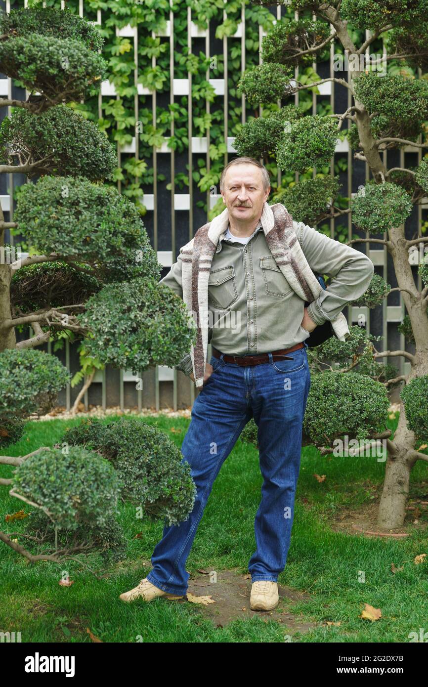 A white-haired fifty-year-old man in jeans and a shirt stands in a city park near a tree Stock Photo