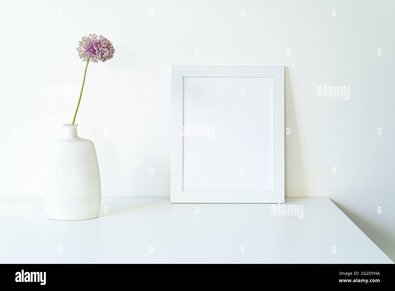 Empty white picture frame mockup. Modern and elegant vase with allium ampeloprasum flower over white table with white background. Minimal and white in Stock Photo