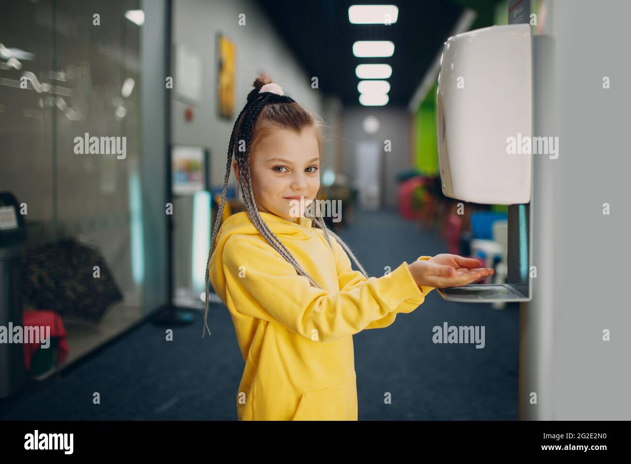 Little girl use automatic sanitizer with alcohol gel dispenser for hands in kindergarten. Antiseptic disinfectant indoor, new normal Coronavirus COVID-19 pandemia concept. Stock Photo