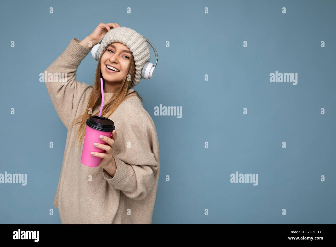 Side-profile photo of Beautiful happy smiling young blonde woman wearing beige winter sweater and hat isolated over blue background drinking beverage Stock Photo