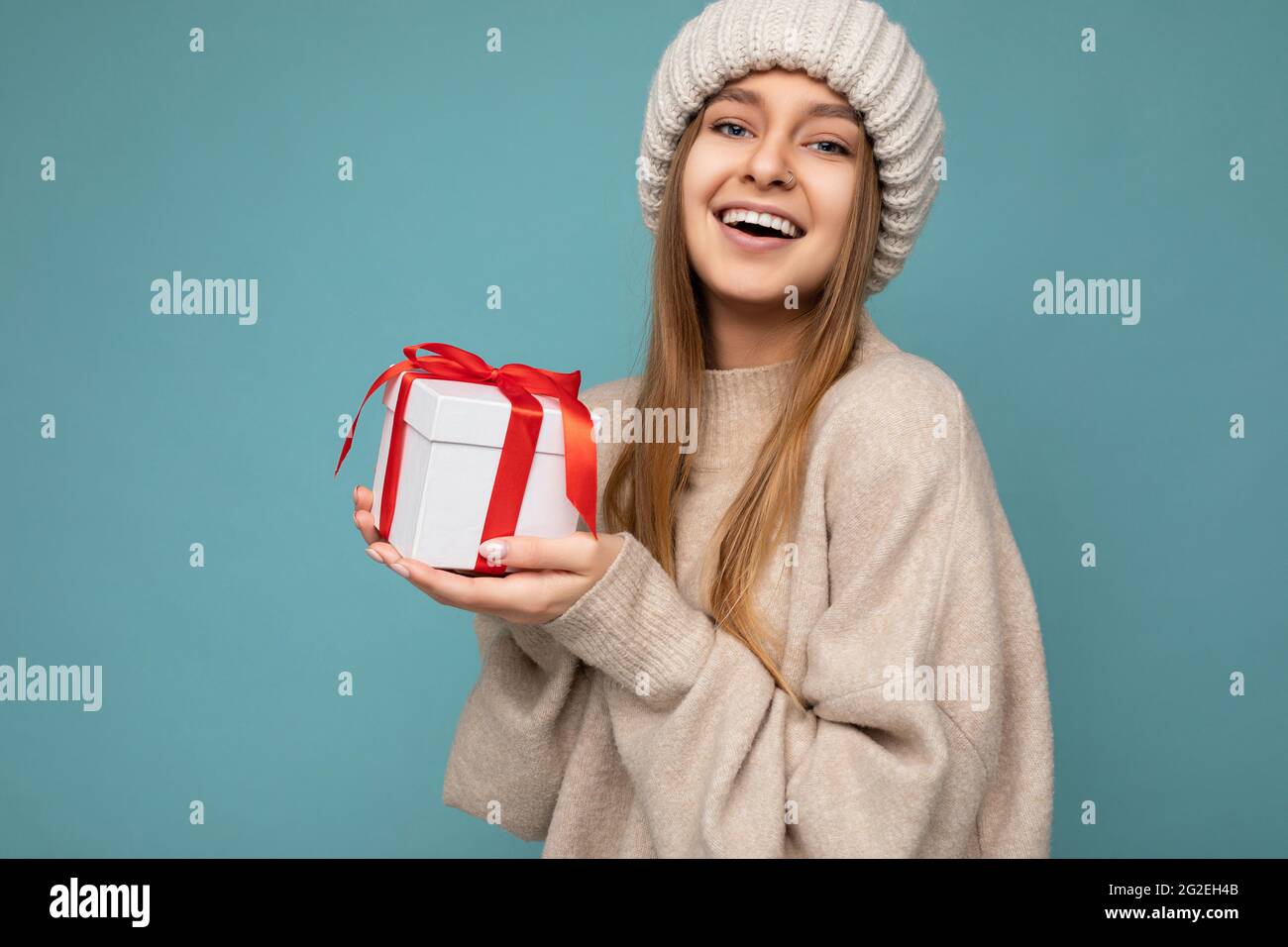 Shot of beautiful positive smiling dark blonde young woman isolated over blue background wall wearing beige warm sweater and beige knitted hat holding Stock Photo