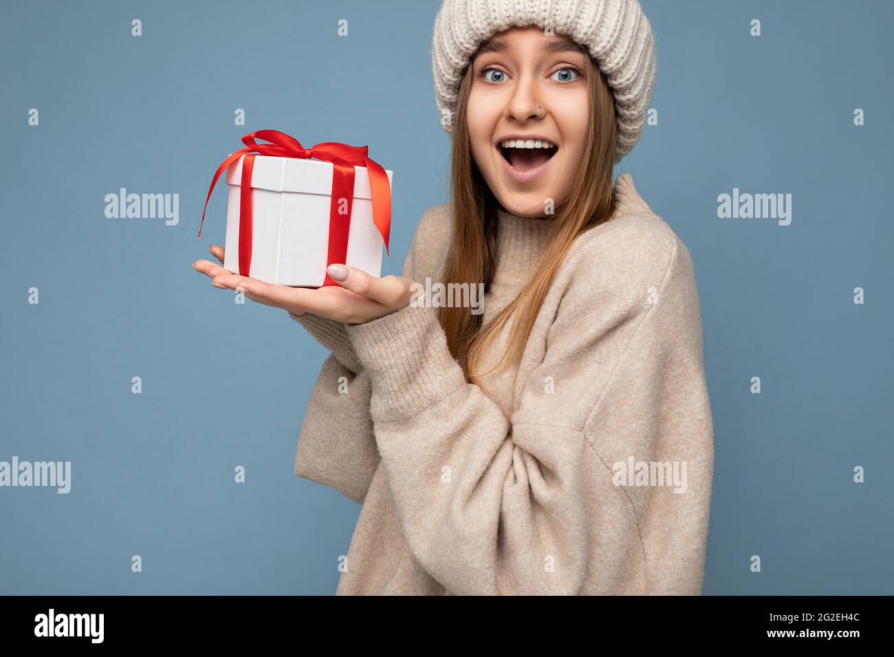 Photo shot of attractive happy surprised dark blonde young woman isolated over blue background wall wearing beige sweater and beige knitted hat Stock Photo