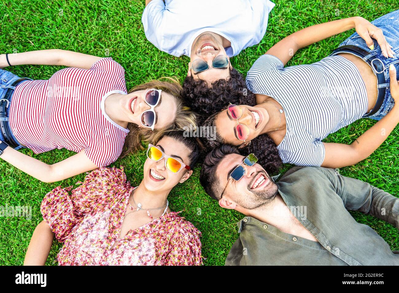 Group of friends lying on park meadow wearing colored trendy sunglasses looking at camera laughing. Happy diverse international people enjoying friend Stock Photo