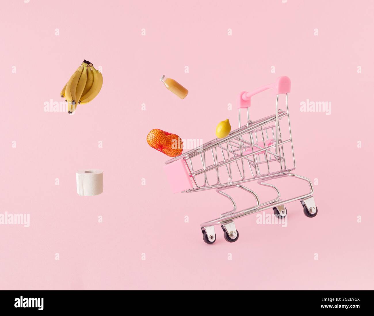 A shopping cart from which different types of food and toilet paper fly on a pink background. Bananas, oranges, juice, lemons fly in several direction Stock Photo