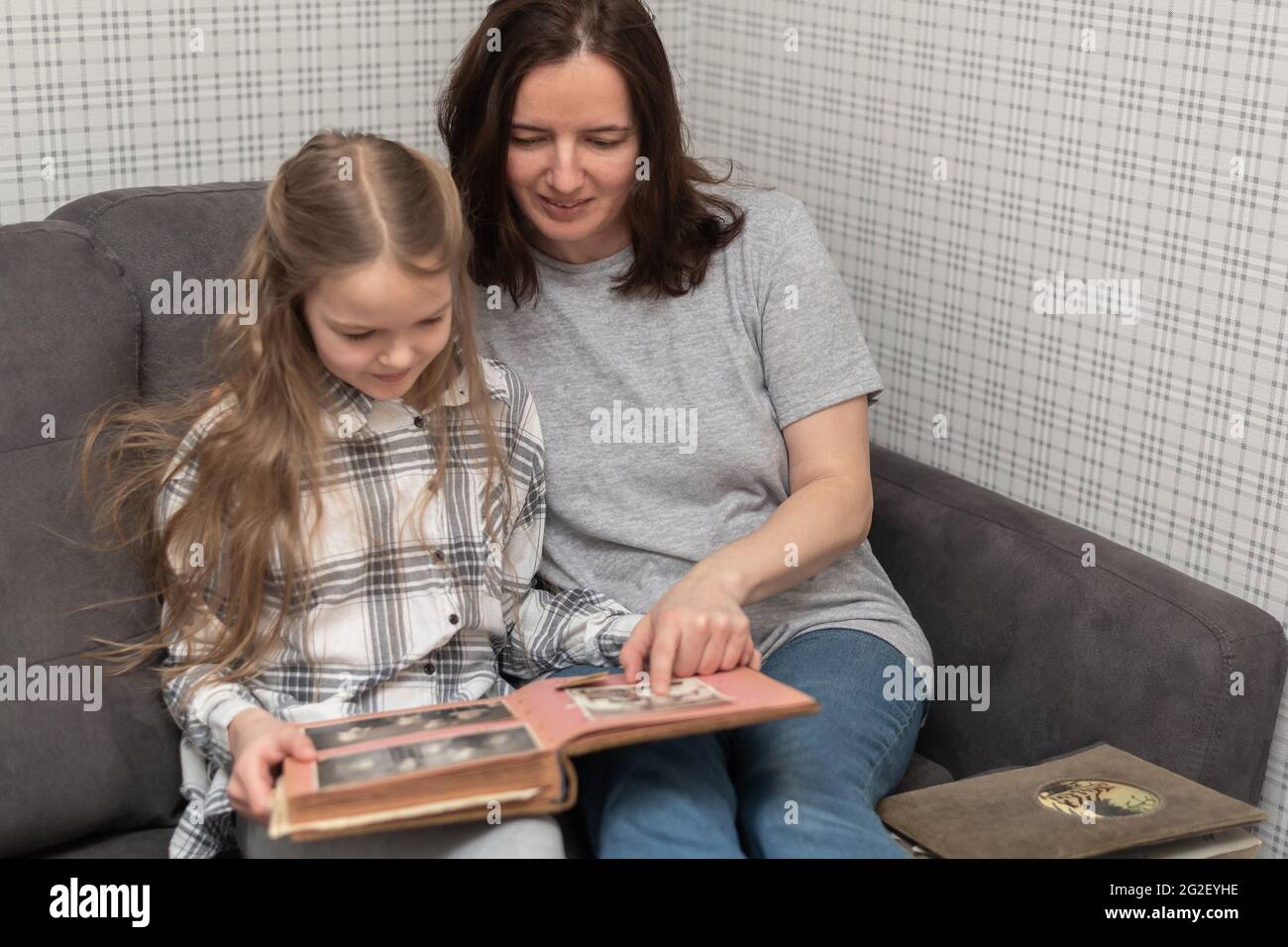 Mom and daughter are sitting on the sofa and look at an old family photo album. Stock Photo