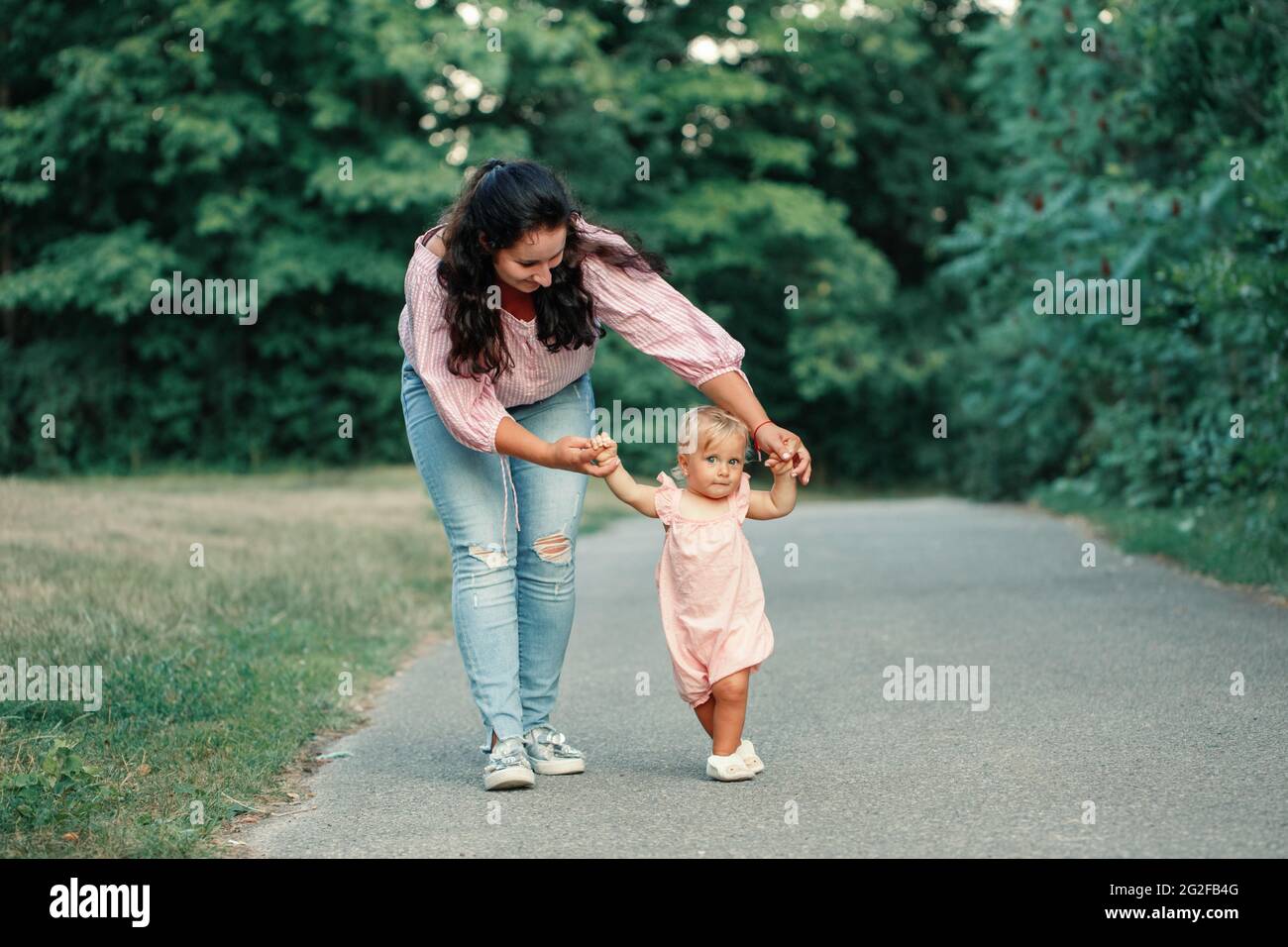 First steps of baby. Cute baby girl learning to walk and holding her mother hand. Mom helping toddler child daughter to go in park outdoors on summer Stock Photo