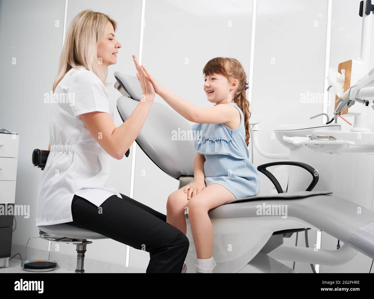 Cute little girl and female dentist giving high five after dental procedure. Smiling child sitting on dental chair and slapping hands with doctor. Concept of pediatric dentistry and dental care. Stock Photo