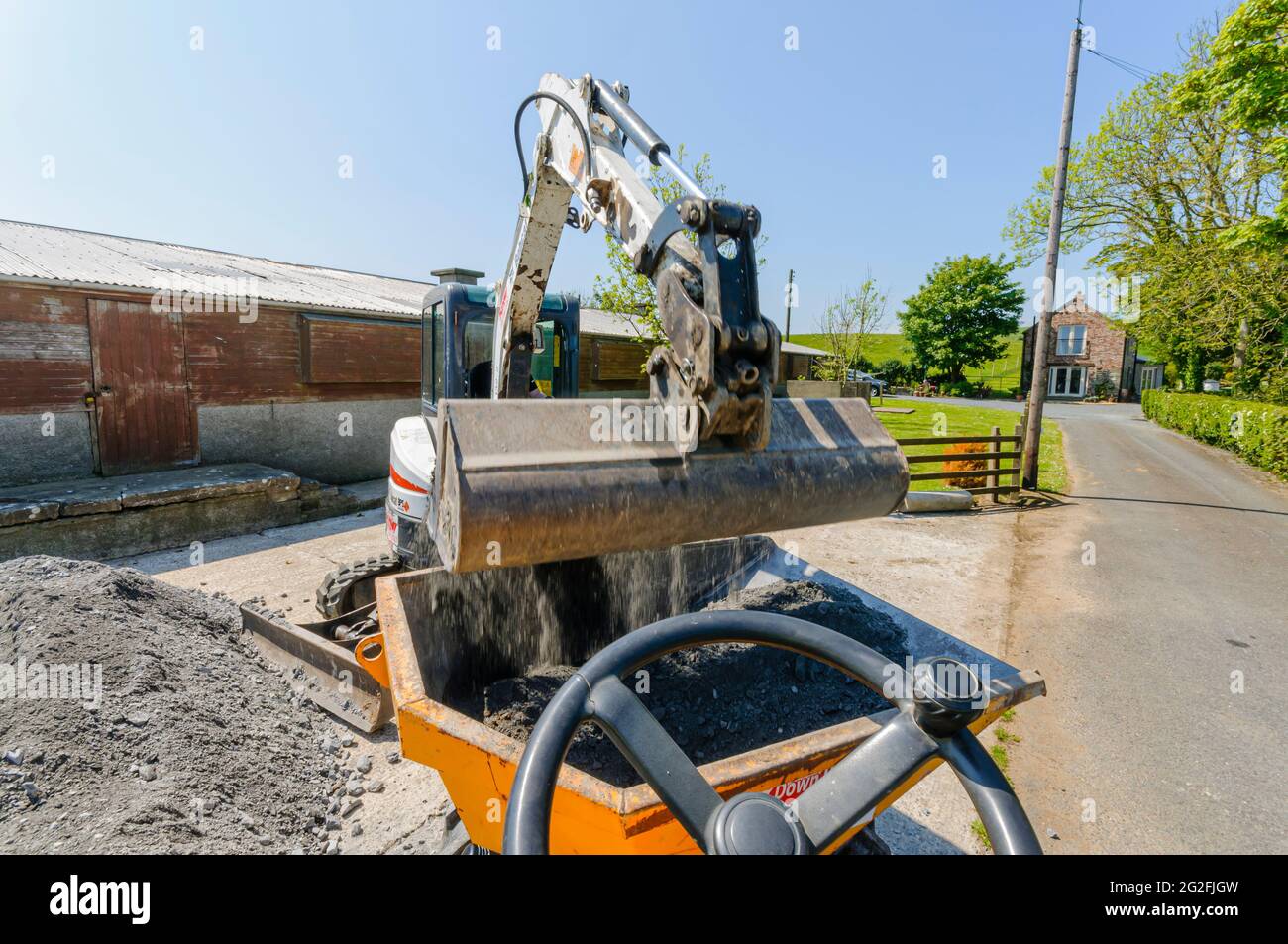 Bobcat E25 mini-digger excavator loads crushed blinding aggregate stones into a yellow dump truck at a building site on a farm. Stock Photo