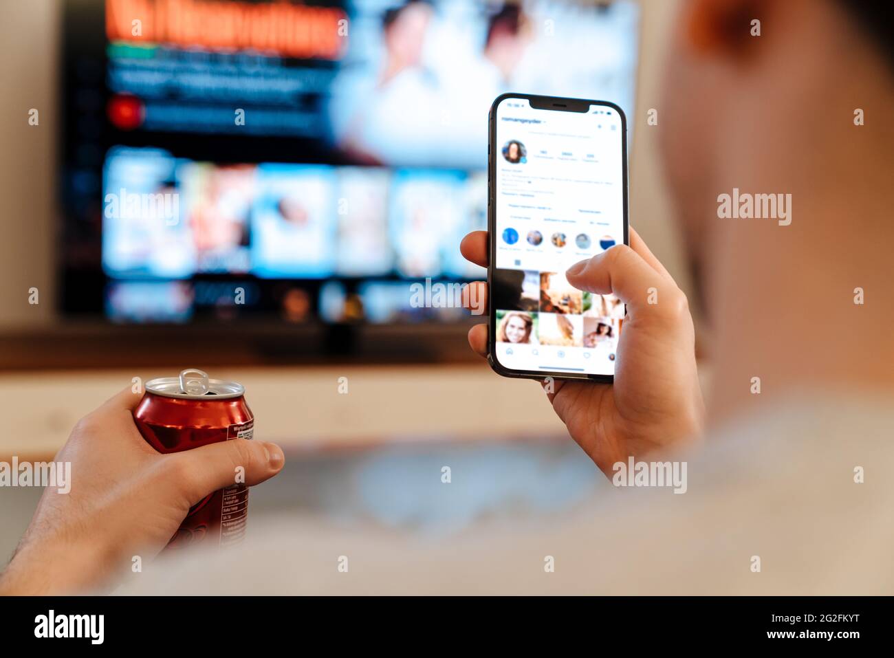 Caucasian guy drinking soda and using mobile phone while watching TV at home Stock Photo