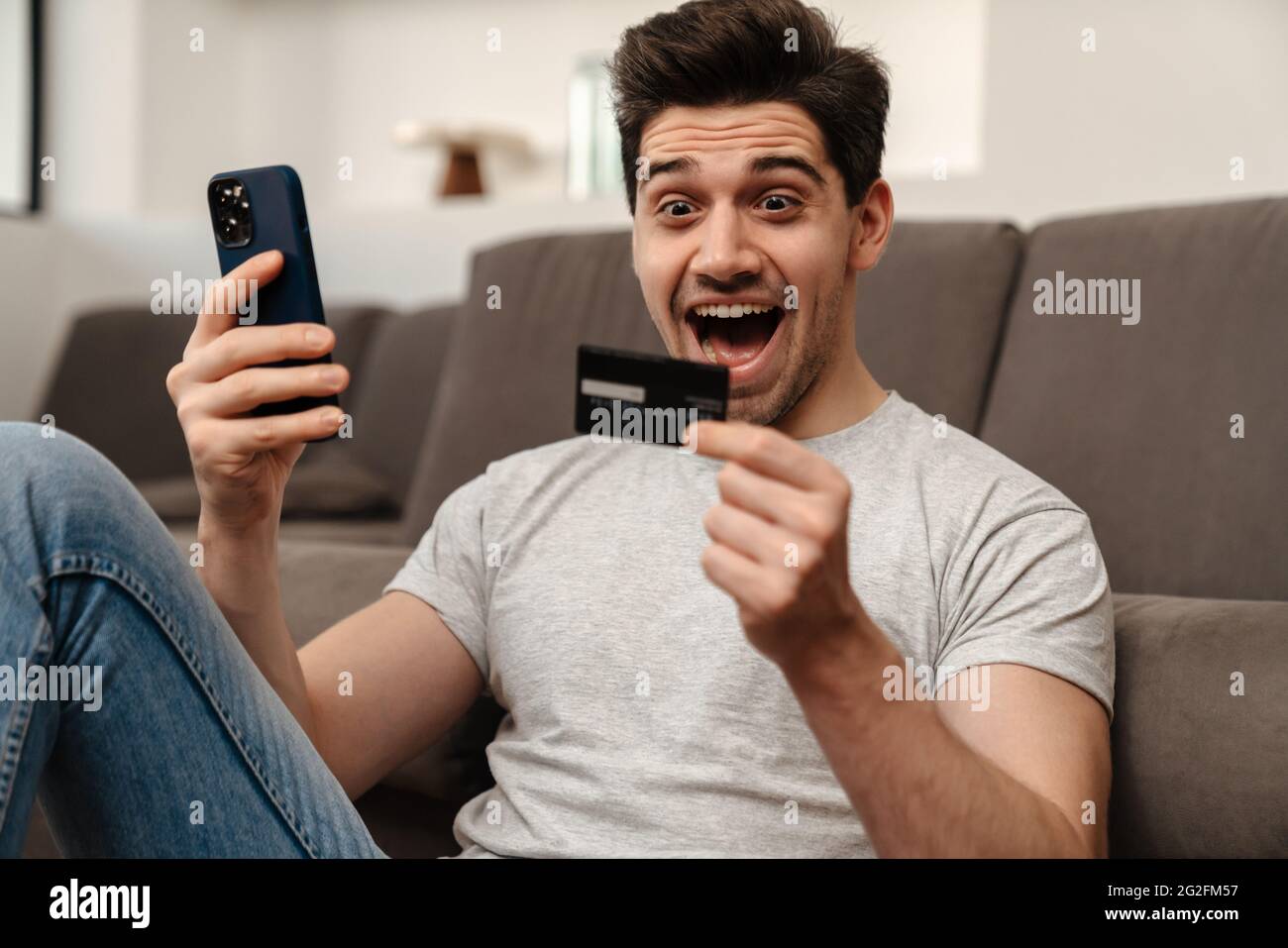 Excited brunette guy using mobile phone and credit card while sitting on floor at home Stock Photo