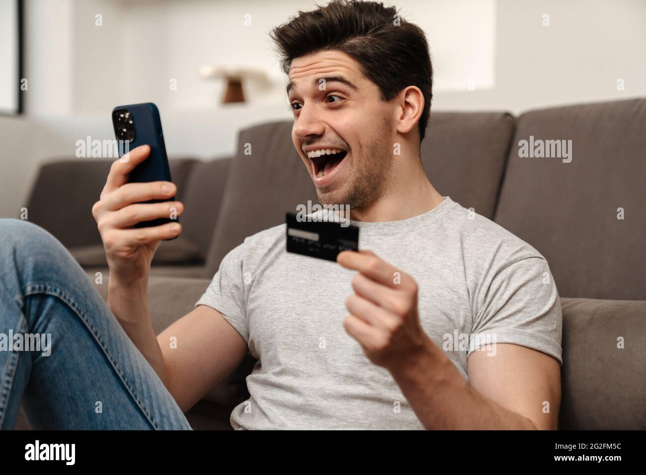 Excited brunette guy using mobile phone and credit card while sitting on floor at home Stock Photo