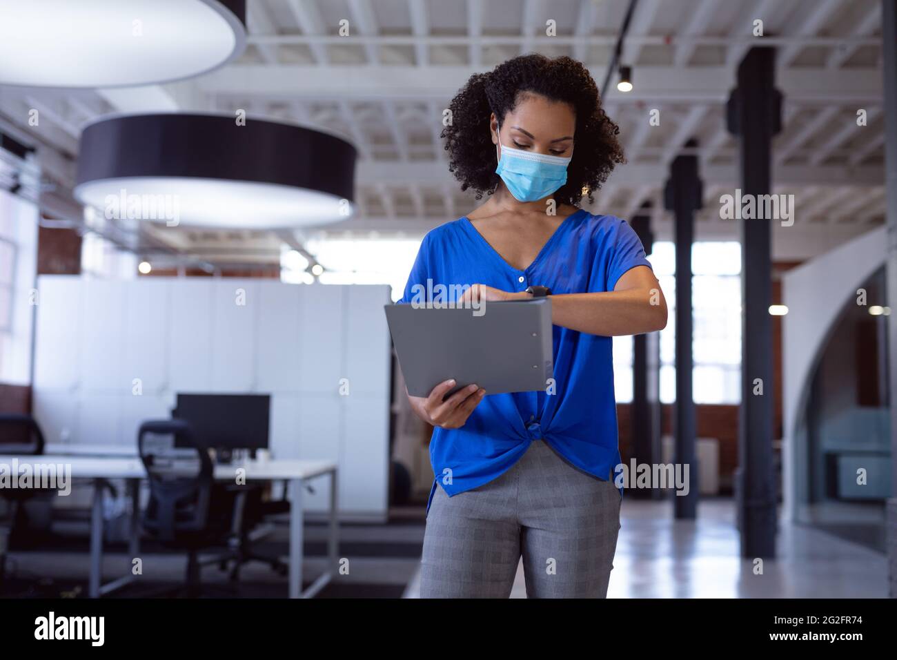 Busy african american businesswoman wearing face mask checking watch in office corridor Stock Photo