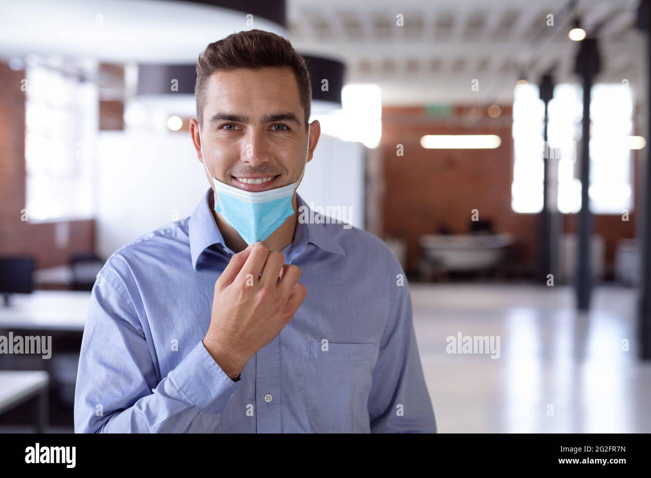 Portrait of happy caucasian businessman lowering face mask and smiling to camera standing in office Stock Photo