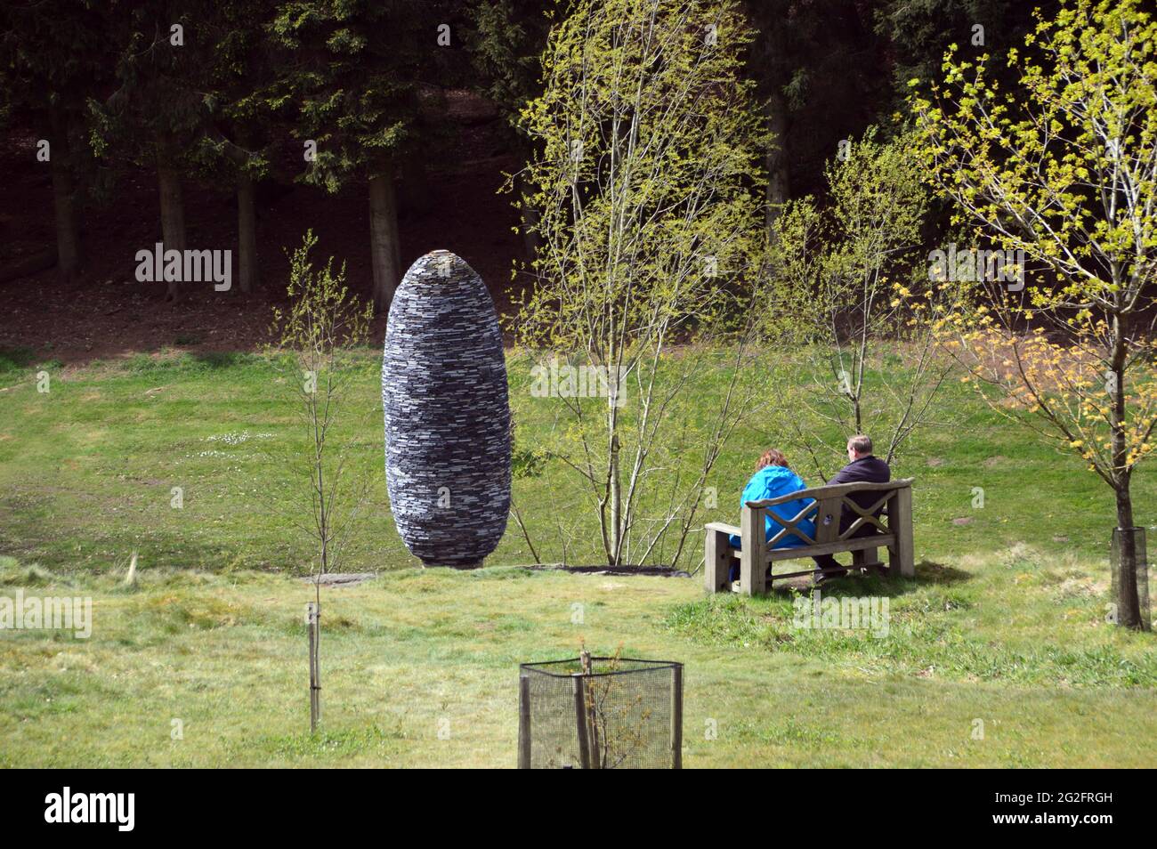 Couple Sitting on Wooden Bench Admiring the Stone 'Fir Cone' Sculpture The Himalayan Garden & Sculpture Park, Grewelthorpe, Ripon, North Yorkshire. Stock Photo