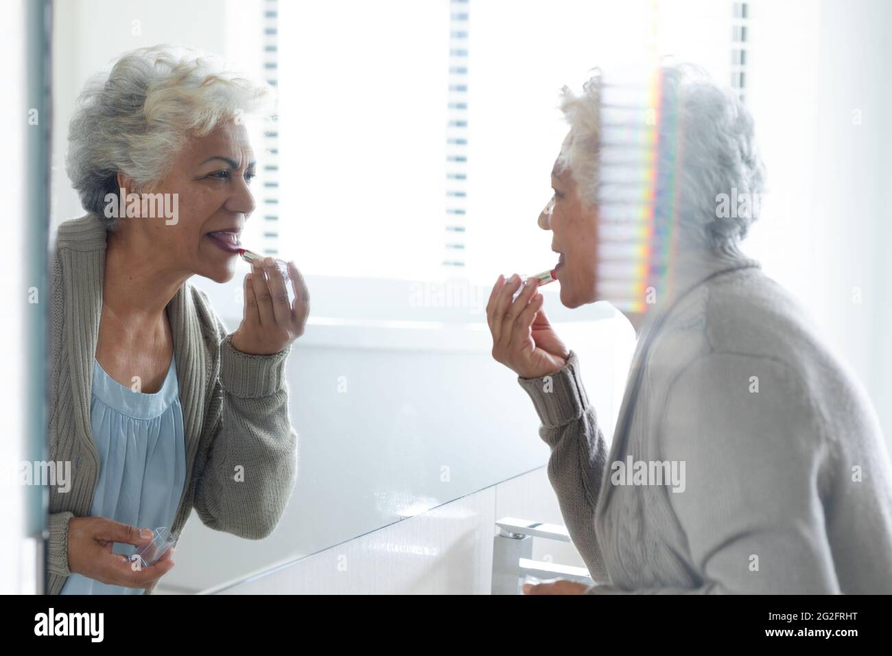 Mixed race senior woman looking at her reflection in mirror wearing lipstick Stock Photo