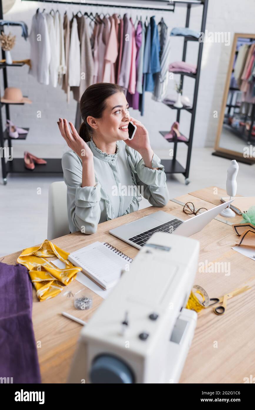 Smiling designer talking on smartphone near laptop and blurred sewing machine in atelier Stock Photo