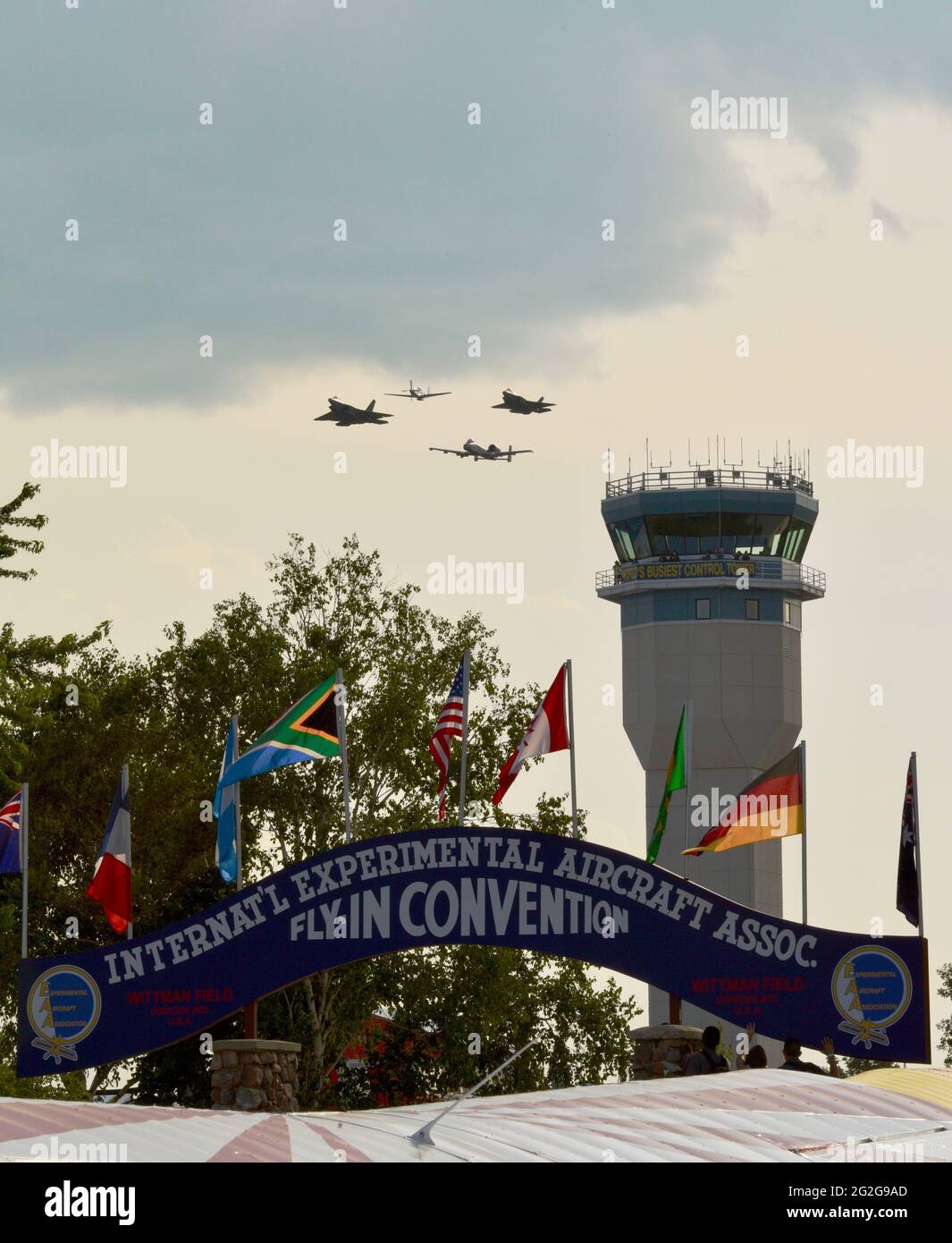 Control tower with F-35, F-22, A-10 fighter jets and P-51 mustang airplane doing fly-by, blue archway at entrance of EAA AirVenture, Oshkosh, WI, USA Stock Photo