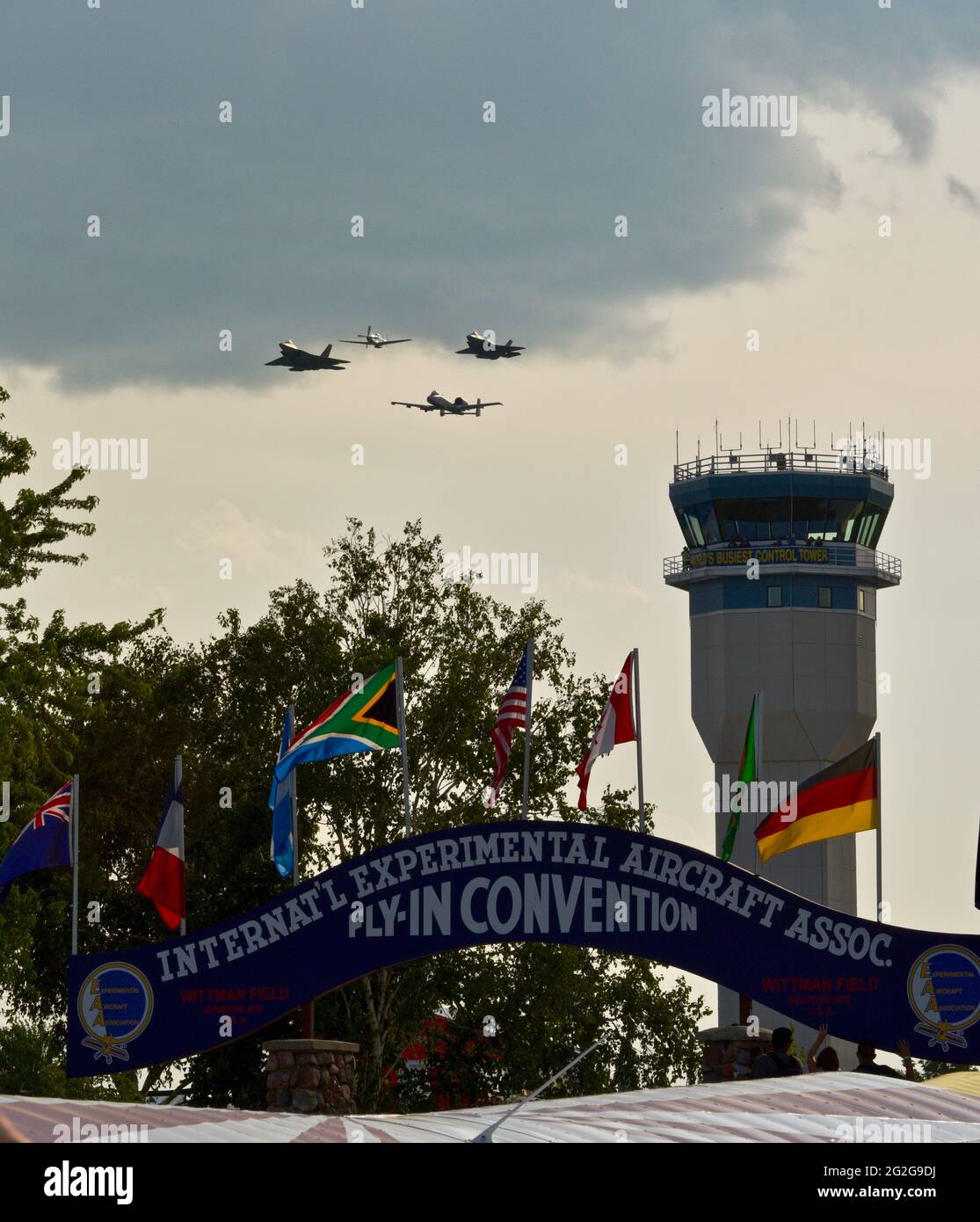 Control tower with F-35, F-22, A-10 fighter jets and P-51 mustang airplane doing fly-by, blue archway at entrance of EAA AirVenture, Oshkosh, WI, USA Stock Photo