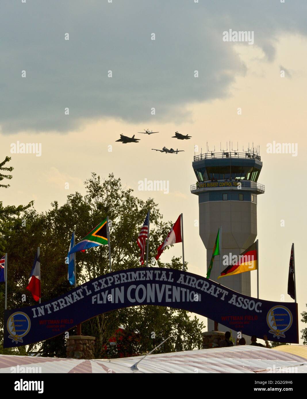 Control tower with F-35, F-22, A-10 fighter jets and P-51 mustang airplane doing fly-by, blue archway at entrance of EAA AirVenture, Oshkosh, WI, USA Stock Photo