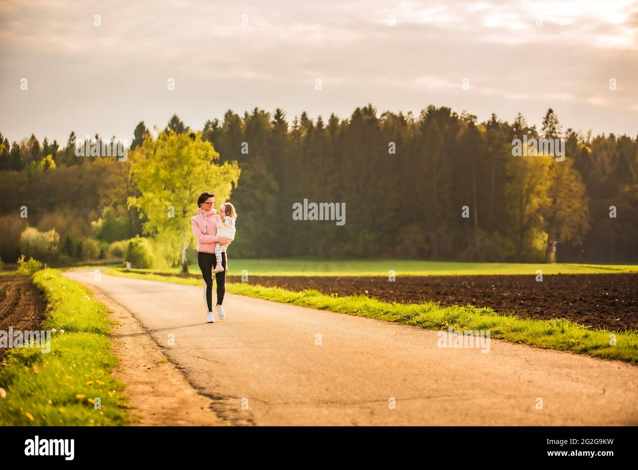 Mother and child walking on countryside road between agricultural fields towards vilage from forest. Stock Photo