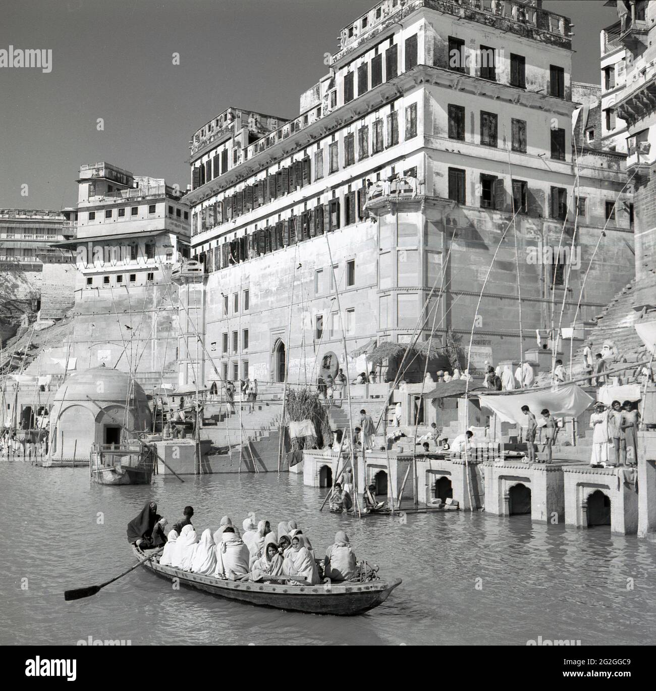 1950s, historical, indian people sitting in a rowing boat on the Ganges river, near the riverbank with its tall buildings in the city of Benares, India. One of the world's largest rivers, it is known as a 'holy river', as the indian people, the majority who are Hindu, consider it sacred and use its waters for religious rituals, many believing that bathing in the waters of the Ganges will cleanse their souls. The Ganges river is therefore an important place of Indian religious activity, culture and history. Stock Photo