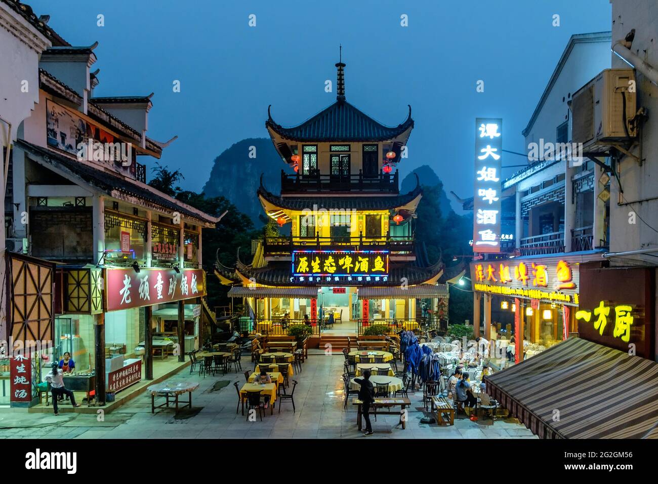 Night view of a square with table and chairs surrounded by illuminated traditional Chinese restaurants against a mountain backdrop, Yangshou, China Stock Photo