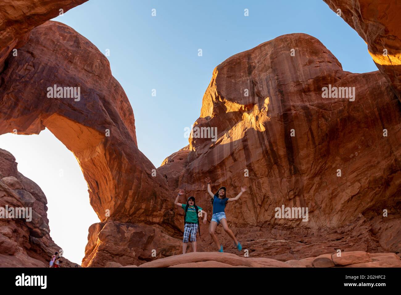 Two happy kids jumping in front of the famous Double Arch - a sandstone formation & popular photo spot with two big arches springing from the same side foundation - noted for front & back spans in Arches National Park, near Moab in Utah, USA Stock Photo