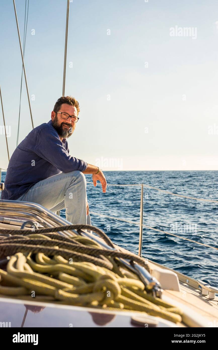Happy mature handsome man sitting and relaxing on sail boat deck enjoying the blue ocean travel adventure alone - free lifestyle and cheerful people on the yacht sailing Stock Photo