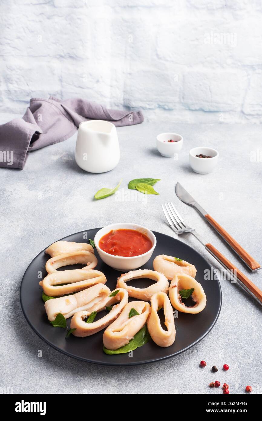 Fried squid rings with tomato sauce and lemon. Black plate, grey concrete background copy space Stock Photo