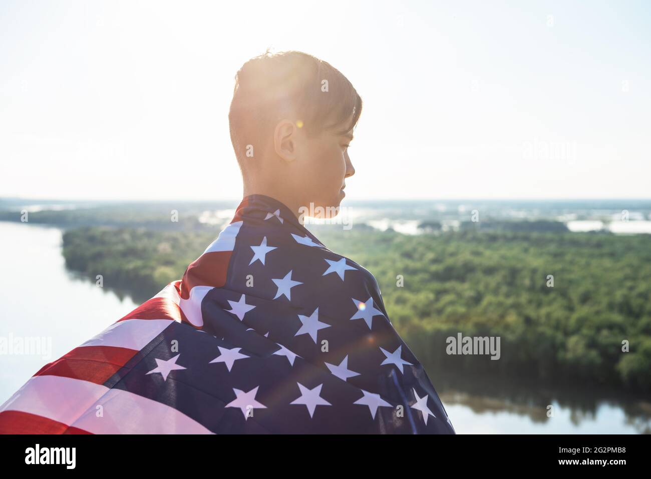 Blonde boy waving national USA flag outdoors over blue sky at the river bank. Beauty summer sunny day. American flag, patriotism, independence day 4th Stock Photo