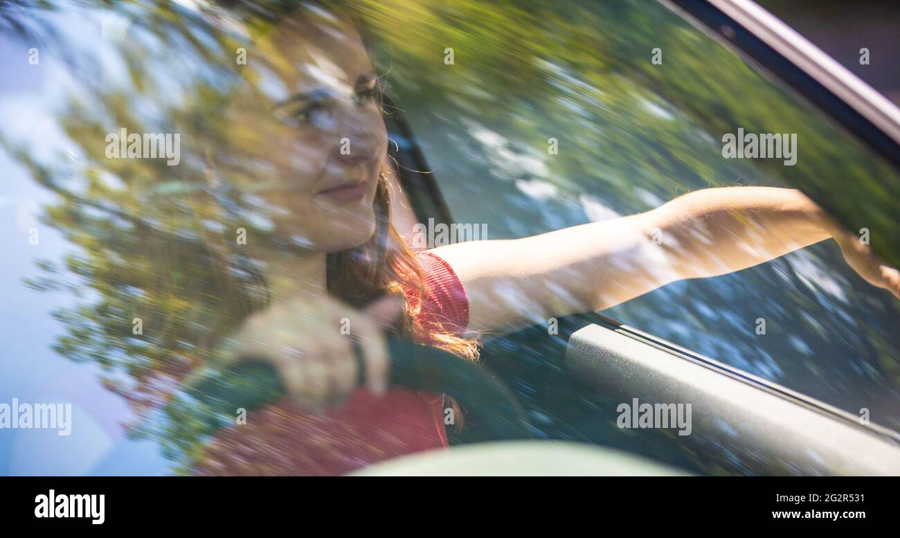 Motion blured image of happy young woman driving a car on a summer road trip travel vacations. Shot trough windshield. Stock Photo