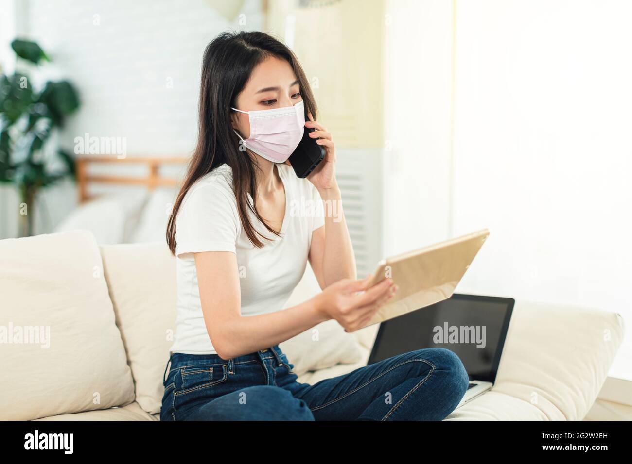 Young woman working at home, wearing a mask to protect herself, sitting on the sofa using a laptop to telecommute, and talking on the phone while watc Stock Photo