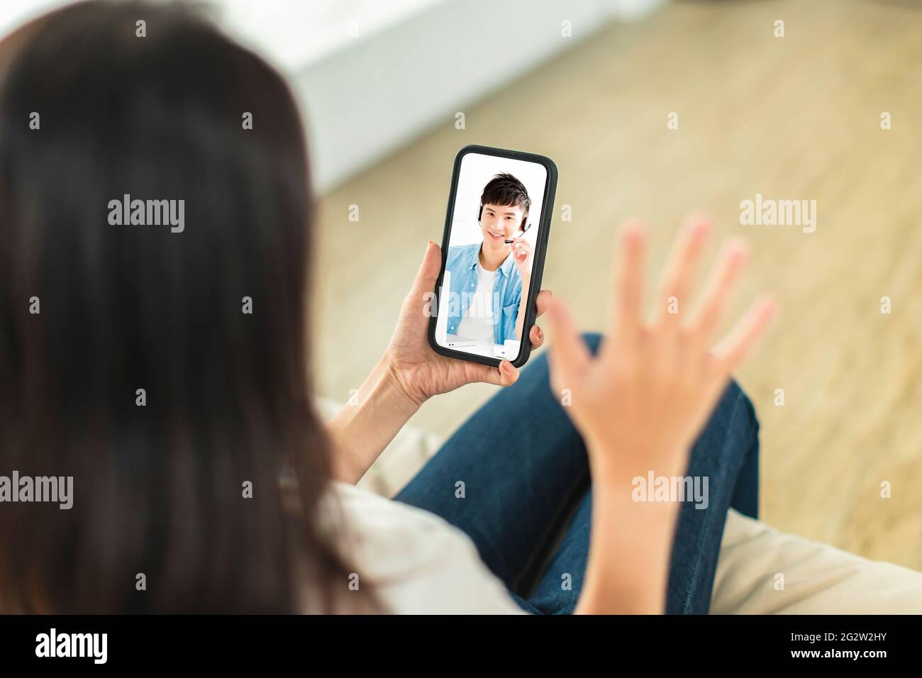 Young woman sitting on sofa at home, holding mobile phone, making video call, discussing with company colleagues in telecommuting Stock Photo