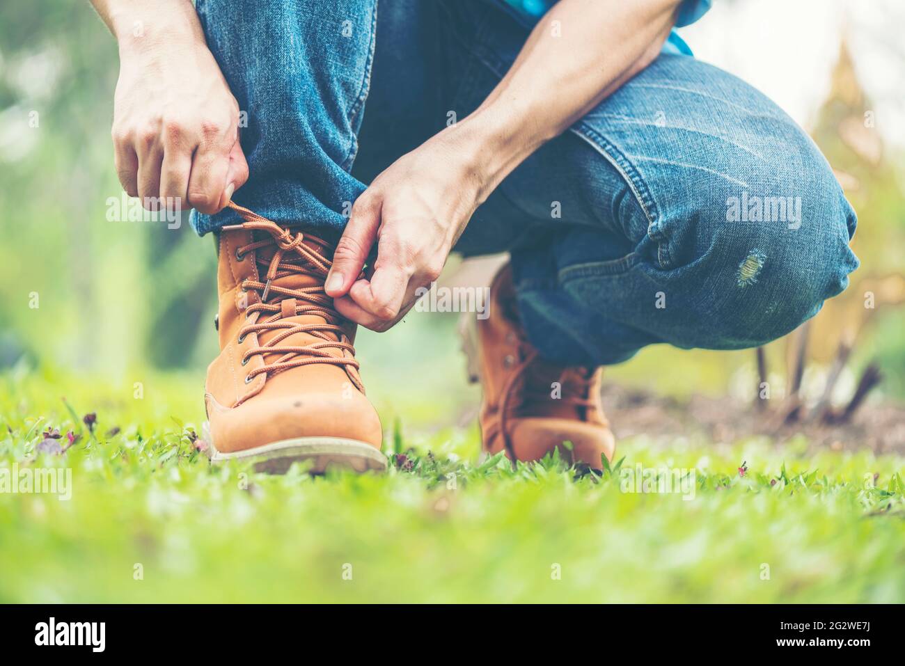 Footwear concept.Handsome man wear jeans knelt down to do up his shoelaces. Preparing before go to party. Brown boots on green grass. close up man han Stock Photo